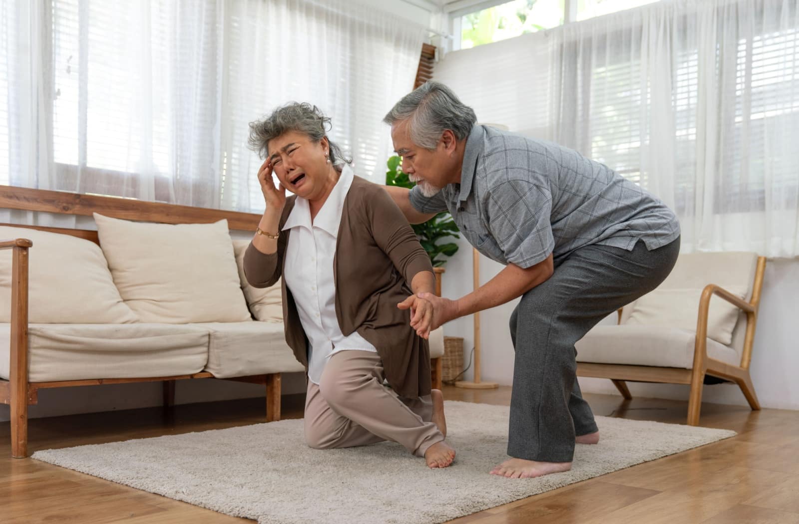 Older adult holding head in pain while another assists them after a fall in a bright senior living room