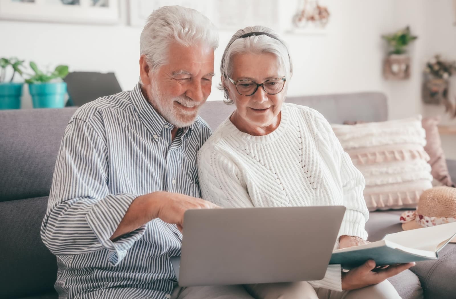 An older adult grins while sitting on the couch beside their spouse and pointing at a laptop screen while researching assisted living