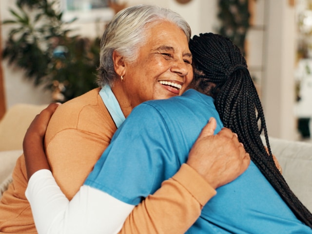 Caregiver in blue scrubs hugs an elderly woman with gray hair who smiles warmly in a home setting, conveying warmth and support.