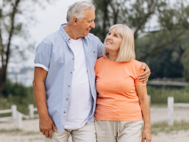 Older couple walking outdoors with arms around each other, smiling as they look at one another.