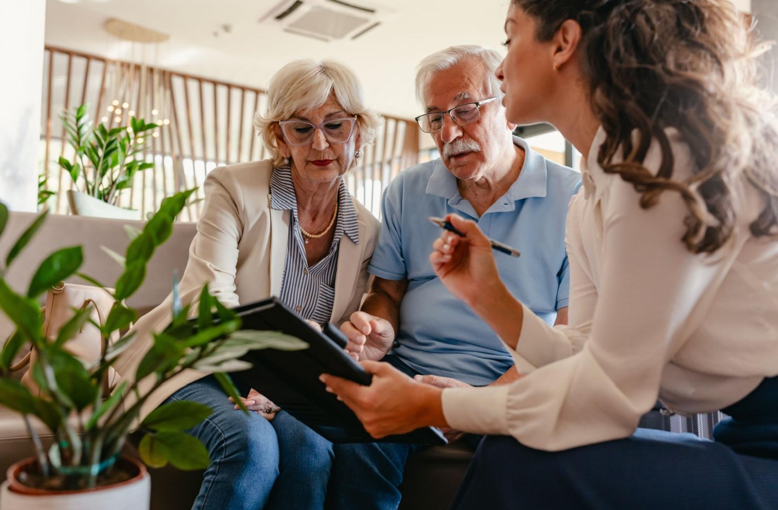 Two older adults look at a tablet while a younger adult speaks with them in a sunlit room.