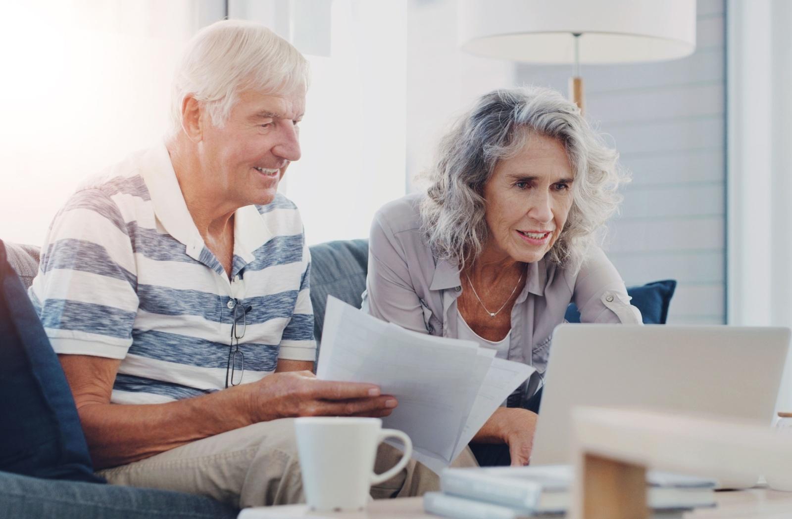 Older couple reviewing documents together on a living room couch with a laptop nearby.