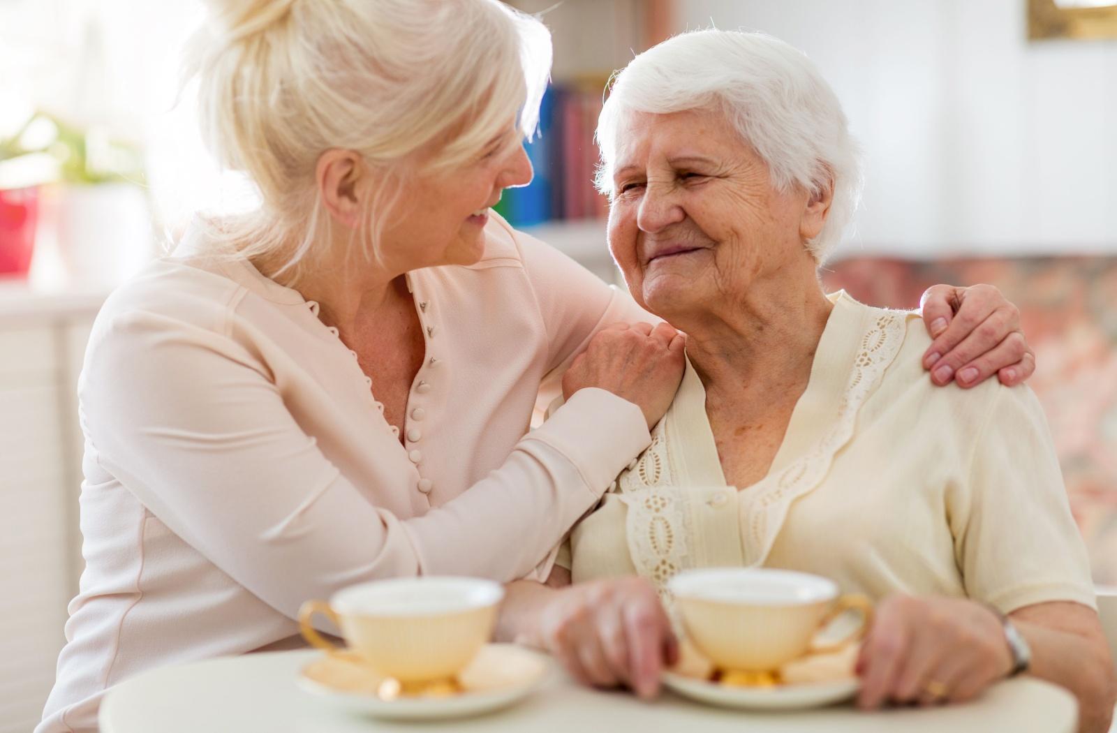 Caregiver smiles at an elderly woman, resting a hand on her shoulder during tea time.